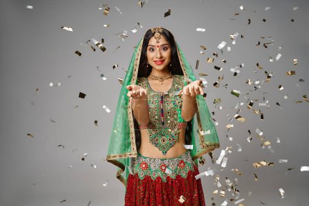 Téléchargez les photos : Belle femme indienne en vêtements nationaux avec bindi montrant les paumes ouvertes et souriant à la caméra - en image libre de droit