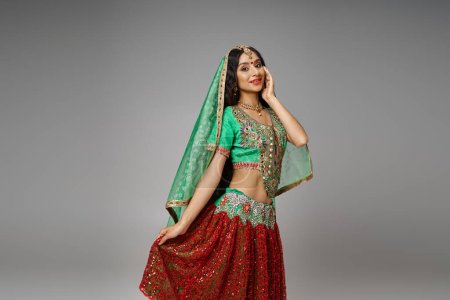 Téléchargez les photos : Joyeuse femme indienne en choli vert touchant l'ourlet de sa jupe rouge posant avec la main sur la joue - en image libre de droit
