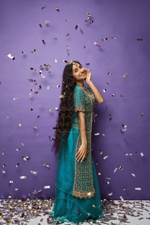 Photo for Attractive cheerful indian woman in blue sari posing under confetti rain on purple background - Royalty Free Image
