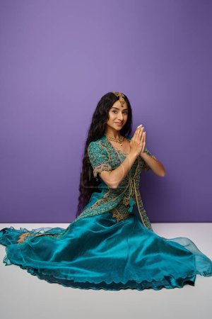 Photo for Vertical shot of young indian woman in blue sari sitting on floor and showing praying gesture - Royalty Free Image