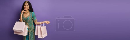Photo for Happy beautiful indian woman in blue sari posing with shopping bags on purple background, banner - Royalty Free Image