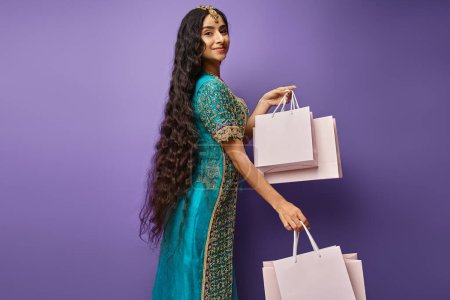 Photo for Pretty jolly indian woman with long hair in blue sari posing with shopping bags on purple backdrop - Royalty Free Image
