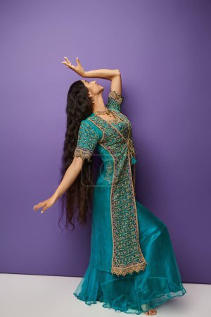 Photo for Young indian woman in blue sari with accessories gesturing while dancing on purple background - Royalty Free Image