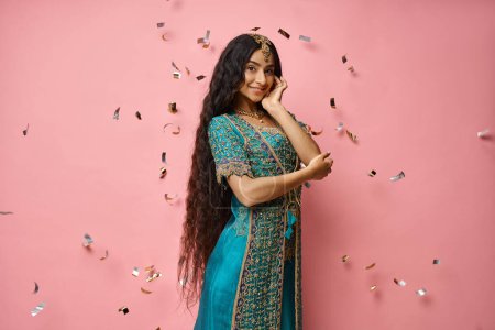 Photo for Cheerful young indian woman in blue sari looking at camera under confetti rain on pink backdrop - Royalty Free Image