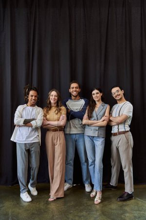smiling multicultural team in stylish casual attire with folded arms near black drape in office