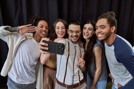 happy man in eyeglasses showing victory sign and taking photo on smartphone with multiethnic team