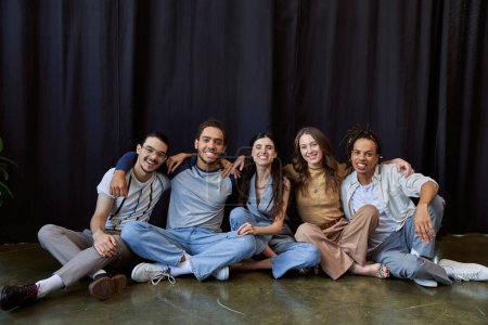 glückliche multiethnische Kollegen, die in der Nähe eines schwarzen Vorhangs im Büro sitzen und in die Kamera lächeln, Gruppenfoto