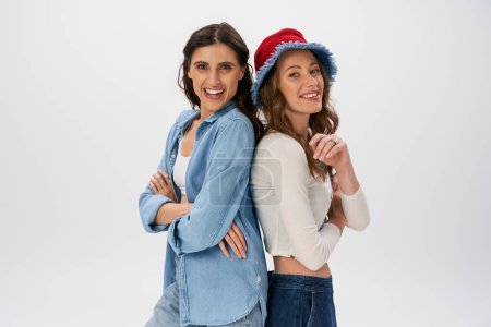 happy women in jeans wear and panama hat posing back to back with folded arms on grey, fashion