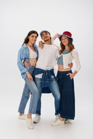 full length of three fashionable friends in trendy outfits and jeans wear posing on grey backdrop