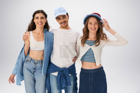 Modische Freunde in lässiger Streetwear umarmen und lächeln vor grauem Hintergrund in die Kamera