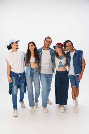 Foto de Diverso grupo de alegres amigos interracial en ropa de calle de moda abrazando y caminando en gris - Imagen libre de derechos