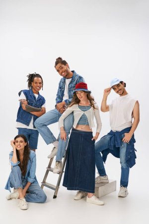 Photo for Smiling multiethnic friends in stylish street wear posing with ladder and looking at camera on grey - Royalty Free Image