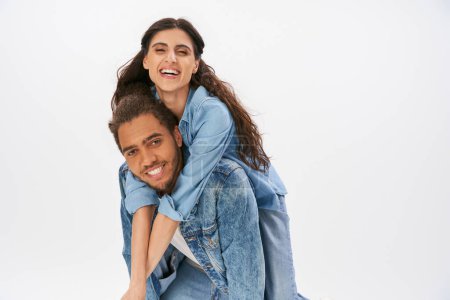 Photo for Smiling african american man in stylish denim clothes piggybacking laughing girlfriend on grey - Royalty Free Image