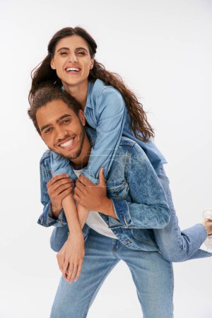 Photo for Young african american man in trendy denim clothes piggybacking laughing girlfriend on grey - Royalty Free Image