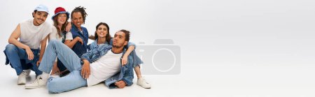 Photo for Trendy african man in denim clothes lying down near happy multiethnic friends posing on grey, banner - Royalty Free Image