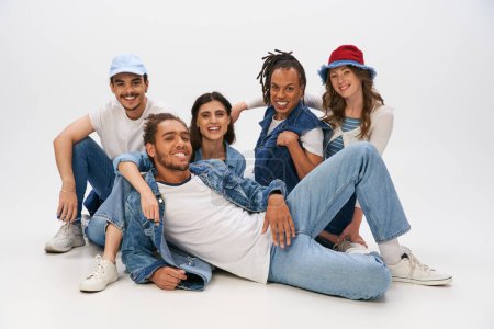 Photo for Cheerful african man in denim wear lying down near interracial friends looking at camera on grey - Royalty Free Image