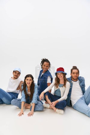 Photo for Happy multiethnic friends in trendy and denim clothes sitting and looking at camera on grey backdrop - Royalty Free Image