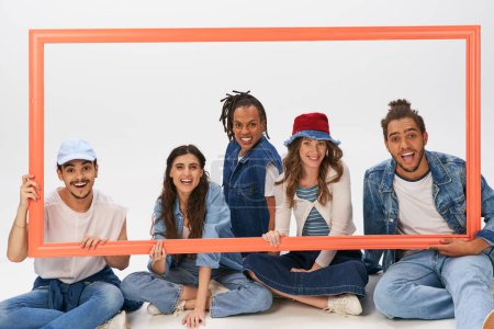 happy and stylish interracial friends sitting and posing with picture frame on grey backdrop