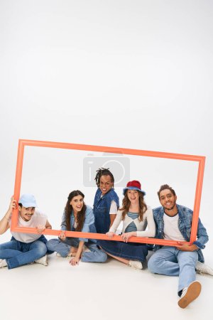 joyful and trendy multiethnic friends sitting and posing with picture frame on grey backdrop