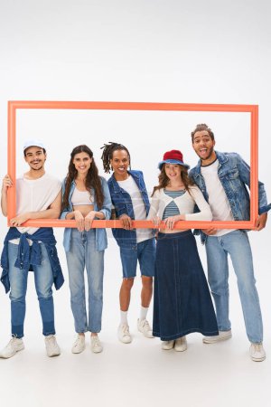 group portrait of multiethnic friends in street wear standing with picture frame on grey backdrop