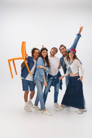 Photo for Happy african american man holding orange chair near trendy interracial friends having fun on grey - Royalty Free Image
