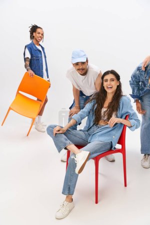 Photo for Laughing woman sitting on chair near fashionable interracial friends on grey, modern fashion - Royalty Free Image