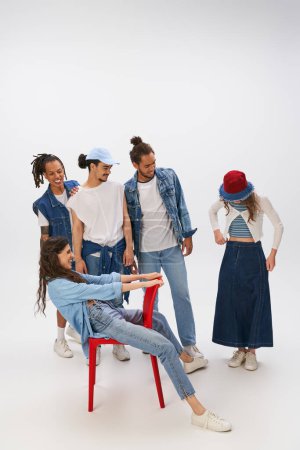 excited woman sitting on red chair near multiethnic friends in stylish street wear on grey backdrop