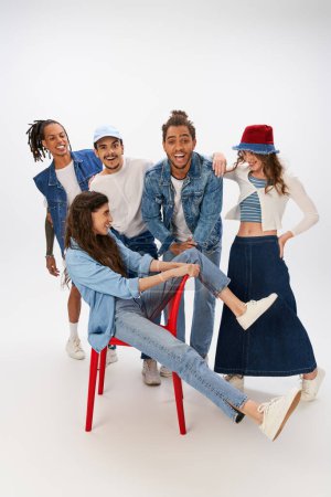 excited woman in denim wear sitting on red chair near trendy interracial friends on grey backdrop
