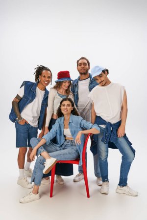 multikulturelle Freunde in stylischer Streetwear lächeln in die Kamera neben einer Frau auf rotem Stuhl auf grau