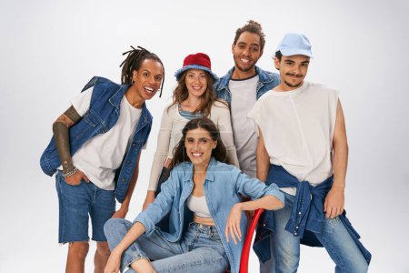 woman in denim wear sitting on red chair and smiling at camera near  interracial friends on grey