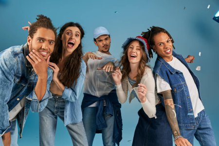 fröhliche multiethnische Freunde in trendiger Freizeitkleidung lächeln unter funkelndem Konfettiregen auf Blau