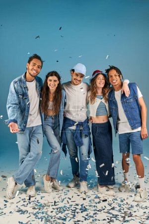 fröhliche multiethnische Freunde in stylischer Streetwear lächeln unter festlichem Konfettiregen auf Blau