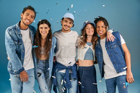glückliche multiethnische Freunde in trendigen lässigen Outfits, die unter Konfettiregen in blau in die Kamera lächeln