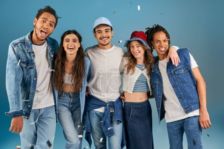 fröhliche interrassische Freunde in trendiger Freizeitkleidung blicken unter Konfettiregen auf blau in die Kamera