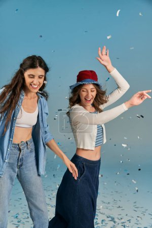excited girlfriends in trendy casual attire having fun under shiny confetti rain on blue backdrop