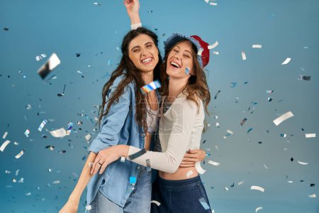 Unbekümmert und trendy Freundinnen umarmen sich unter funkelndem Konfettiregen auf blauem Hintergrund, Party-Time