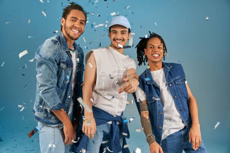 fröhliche multikulturelle Freunde in stylischer Jeans lächeln in die Kamera unter glänzendem Konfetti auf Blau