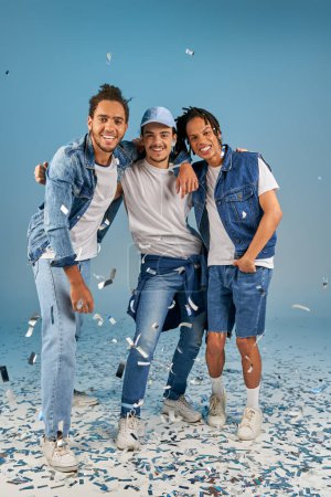fröhliche interrassische Freunde in stylischer Jeanskleidung umarmen sich unter glänzendem Konfettiregen auf Blau