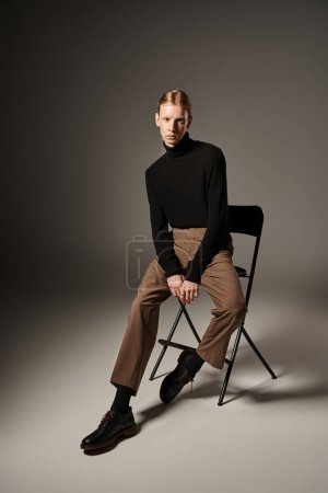 attraktive nicht binäre Person in schwarzem Rollkragen und brauner Hose auf schwarzem Stuhl sitzend, Mode