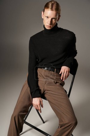good looking young non binary person with ponytail sitting on black chair and looking at camera