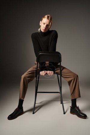 attraktive nicht binäre Person ohne Rollkragen und braune Hose auf schwarzem Stuhl sitzend, Mode