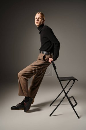 Foto de Modelo no binario de buen aspecto en cuello alto negro apoyado en silla negra y mirando a la cámara - Imagen libre de derechos