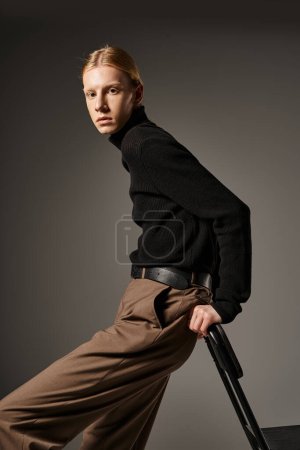 attractive young non binary person in black turtleneck leaning on black chair, looking at camera