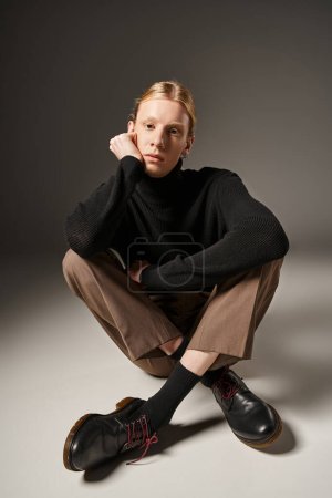 Foto de Buen aspecto joven modelo no binario sentado en el suelo con las piernas cruzadas y la mano cerca de la cara, la moda - Imagen libre de derechos