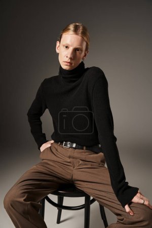 good looking young non binary person in stylish turtleneck sitting on black chair looking at camera