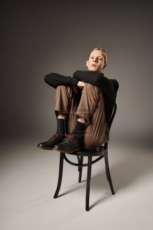 Foto de Plano vertical de joven persona no binaria en cuello alto negro sentado en la silla y mirando a la cámara - Imagen libre de derechos