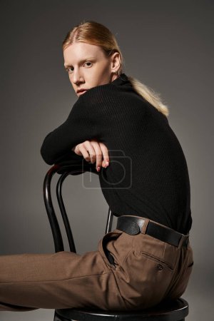 vertical shot of stylish non binary model in trendy outfit sitting on chair and looking at camera