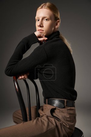 Foto de Plano vertical de joven modelo no binario posando en silla negra con la mano bajo la barbilla mirando a la cámara - Imagen libre de derechos