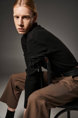 junge nicht binäre Person in stylischem schwarzen Rollkragen mit Pferdeschwanz posiert auf Stuhl, Modekonzept