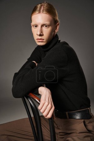 Foto de Modelo no binario de buen aspecto en traje elegante con el pelo rojo sentado en la silla y mirando a la cámara - Imagen libre de derechos